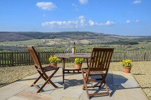 Um pátio com duas cadeiras de madeira e uma mesa, ladeado por vasos com flores amarelas, com vista para uma paisagem deslumbrante.