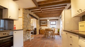 Cuina amb paret de pedra, bigues de fusta, zona de menjador amb taula i cadires, i nevera amb la inscripció "BREAD".