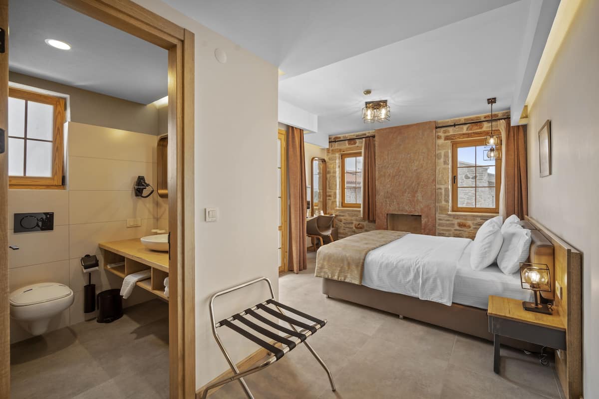 A bedroom with a bed, bedside table, and a chair. There is a bathroom with a toilet, sink, and shower. The room has a stone wall and a window.