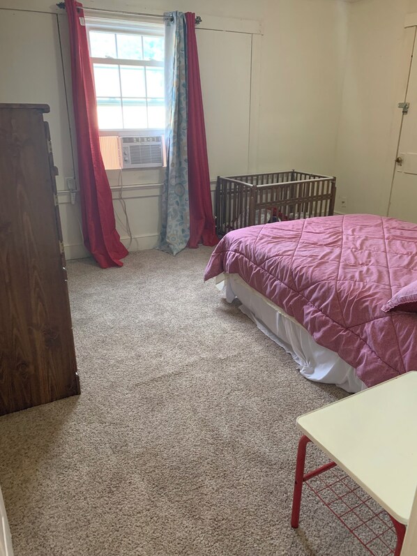 Un dormitorio con una cuna, una cama con colcha rosa, un aparador de madera y una ventana con cortinas.