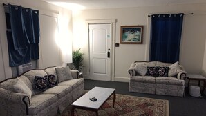 Una sala de estar con dos sofás de estampado floral, una mesita, un tapete decorado y una puerta con un cuadro encima.