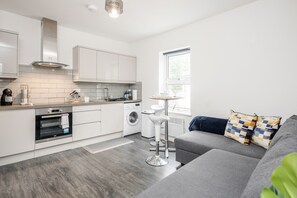 A modern kitchen with white cabinetry, a built-in oven, and a washing machine. There is a grey sofa with patterned cushions and a small table with stools. - 1BR Victorian House Near Bedford City Centre (Bedford)