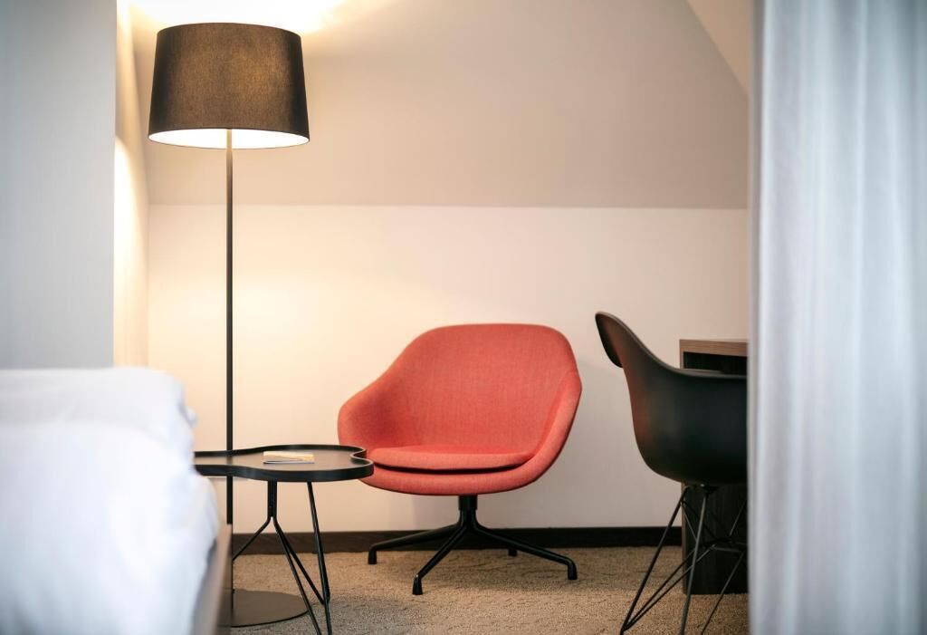 A hotel room with a bed, a red chair, a black chair, a small table, and a floor lamp.