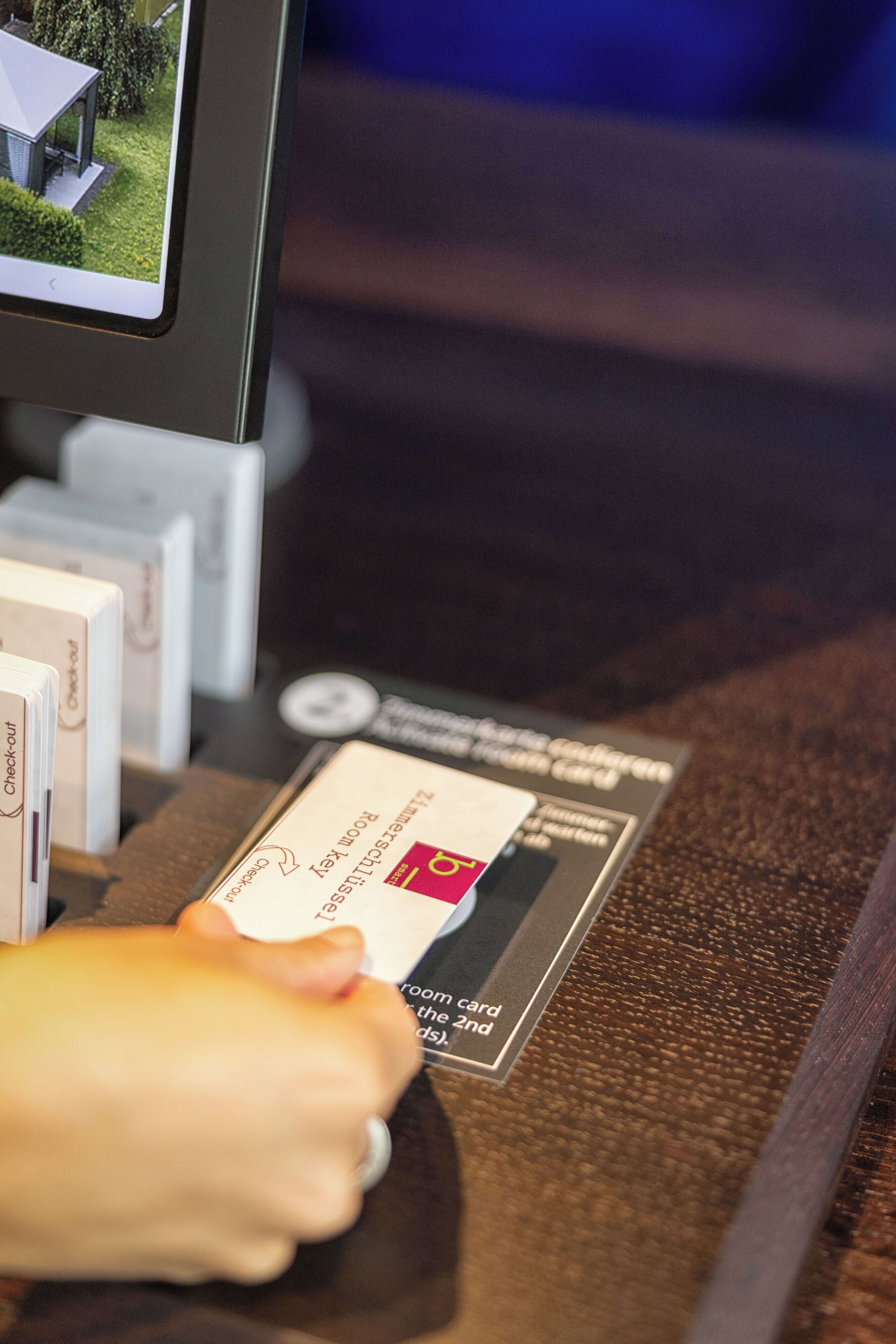 Une main tenant une carte de chambre avec un symbole indiquant le départ, un écran affichant une image d’un bâtiment et un comptoir où sont exposés divers articles.