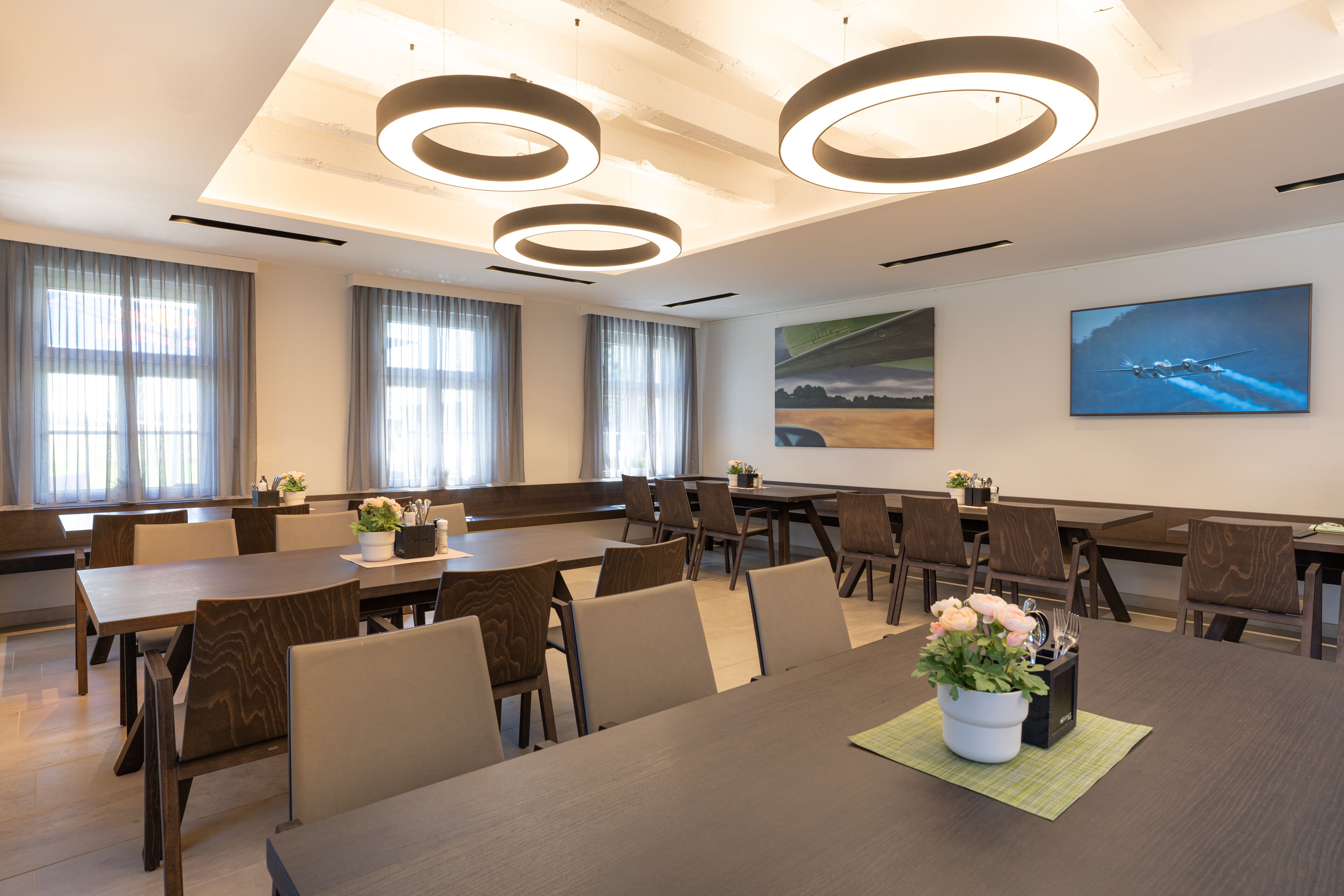 Une salle de conférence dotée de plusieurs tables et chaises, de grandes fenêtres avec des rideaux et d’un tableau représentant un avion accroché au mur.