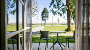 Un patio al aire libre con una mesa redonda y dos sillas, un sendero que lleva a un campo deportivo y árboles al fondo.