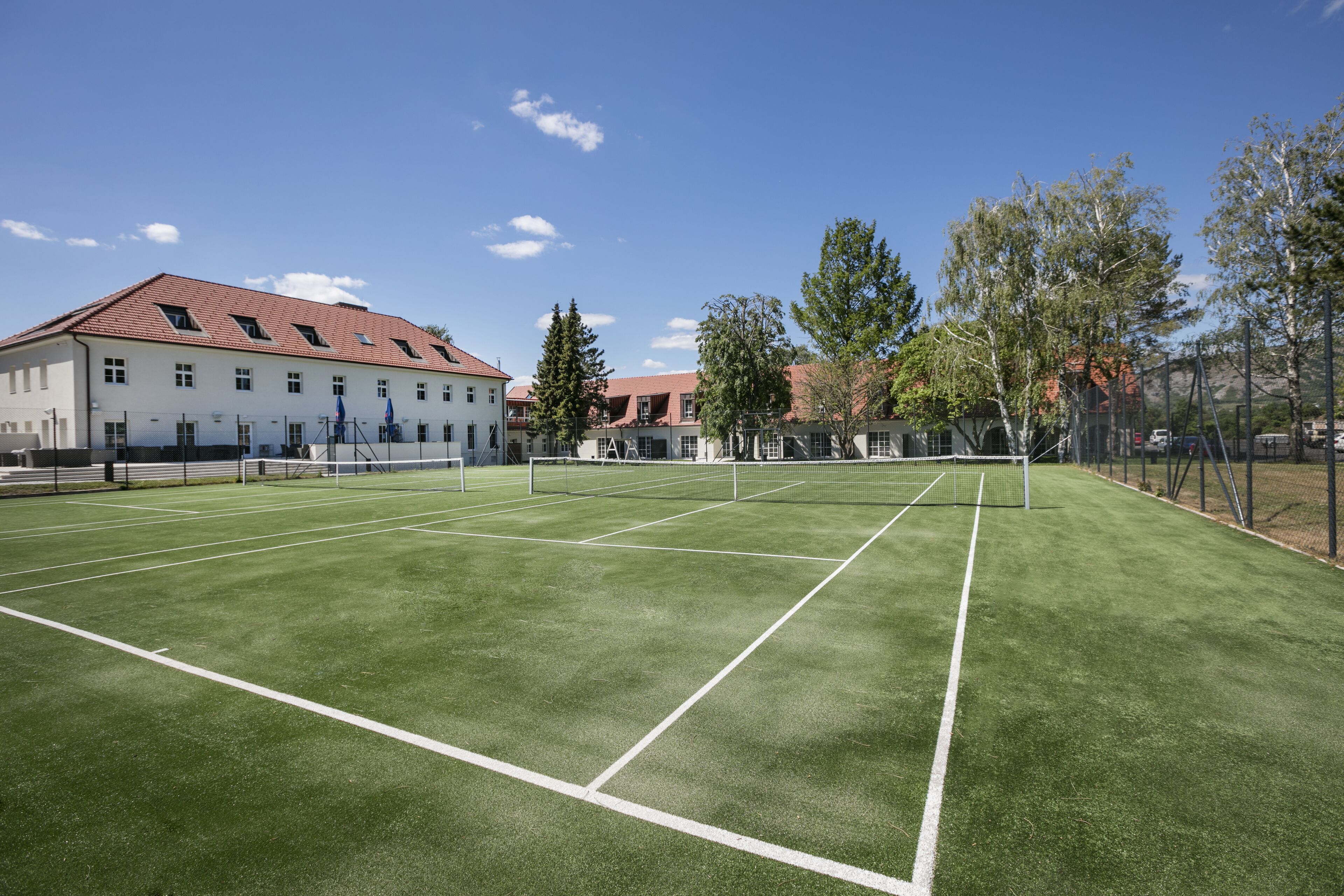 Un court de tennis avec un filet, entouré d’arbres et bordé d’un bâtiment dont le toit est rouge.