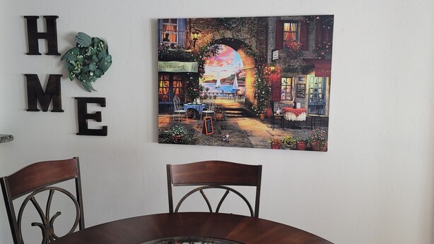 A dining area with a table and chairs, a wall-mounted sign reading "HOME", and a painting of a restaurant scene. - New house with fresh design available in Olsen TH. (Tucson)