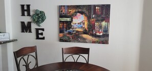 A dining area with a table and chairs, a wall-mounted sign reading "HOME", and a painting of a restaurant scene.