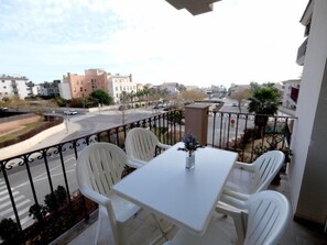 A balcony with a table and chairs, a vase with flowers, and a view of a street and buildings. - Golden Beach by Interhome (St Carles de la Ràpita)