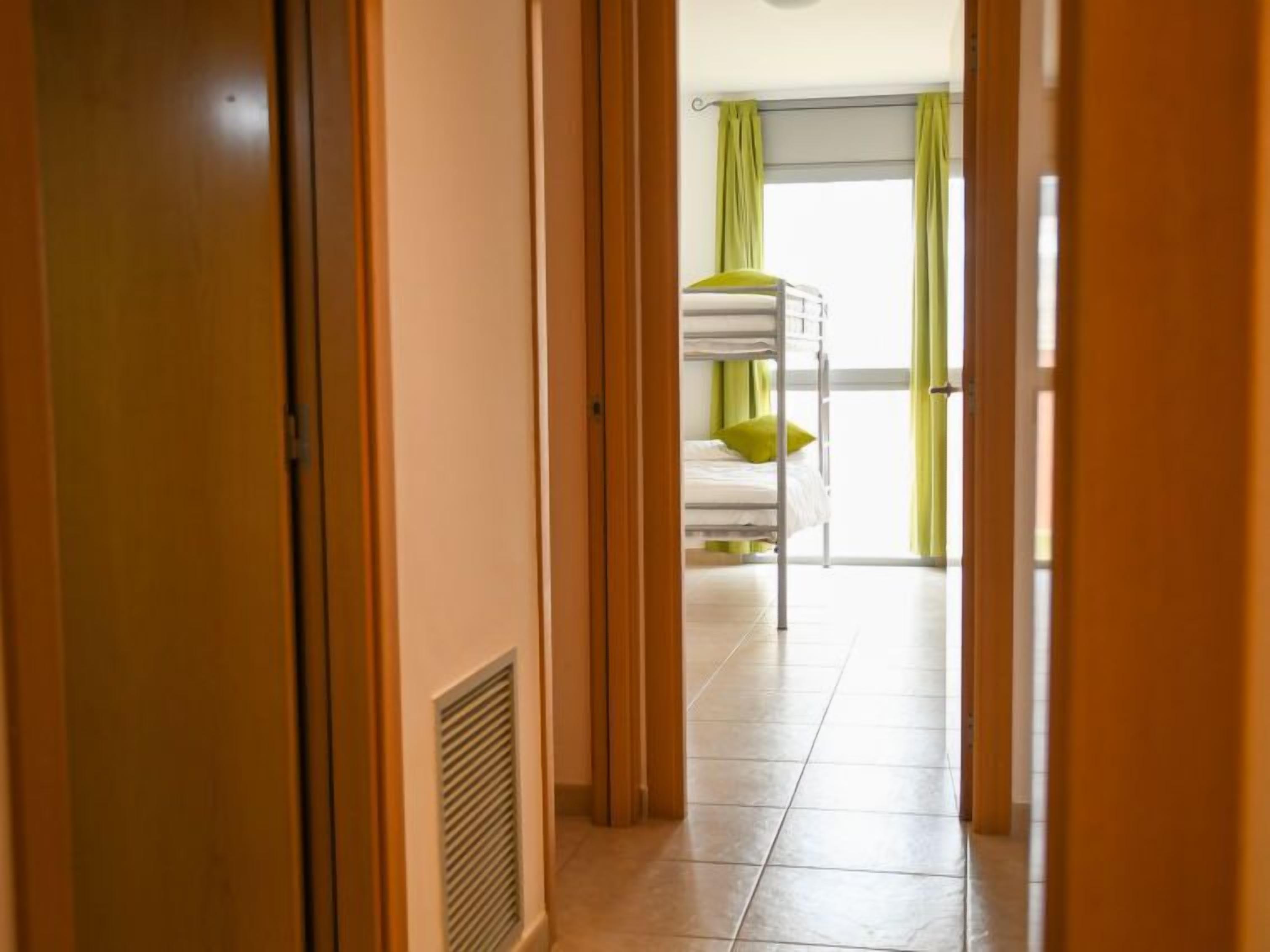 Een gang die uitkomt in een kamer met stapelbedden en een raam met groene gordijnen.