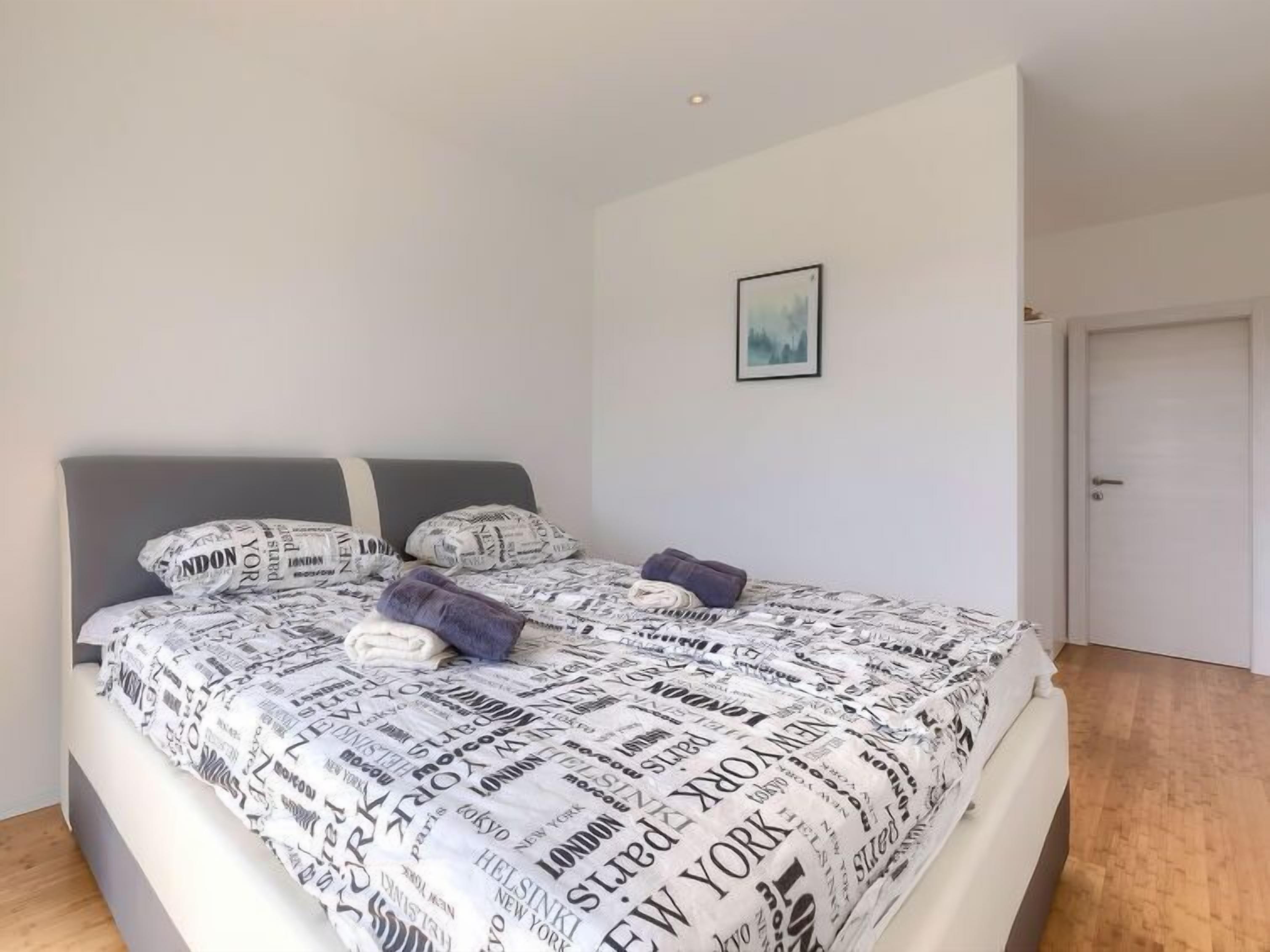 A bedroom with a bed covered in a newspaper print bedspread, a grey headboard, and a framed picture on the wall.