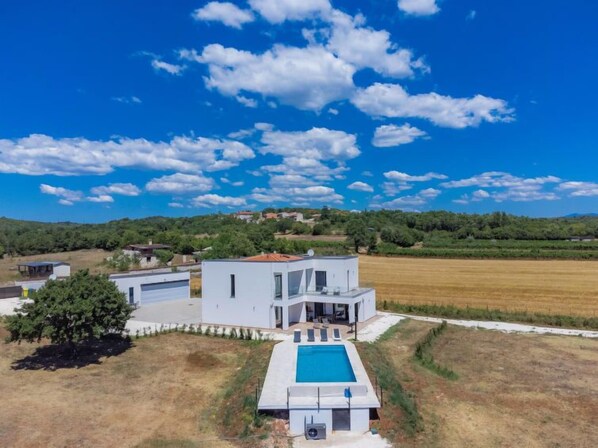 A modern house with a pool, surrounded by a field and trees, under a clear sky.