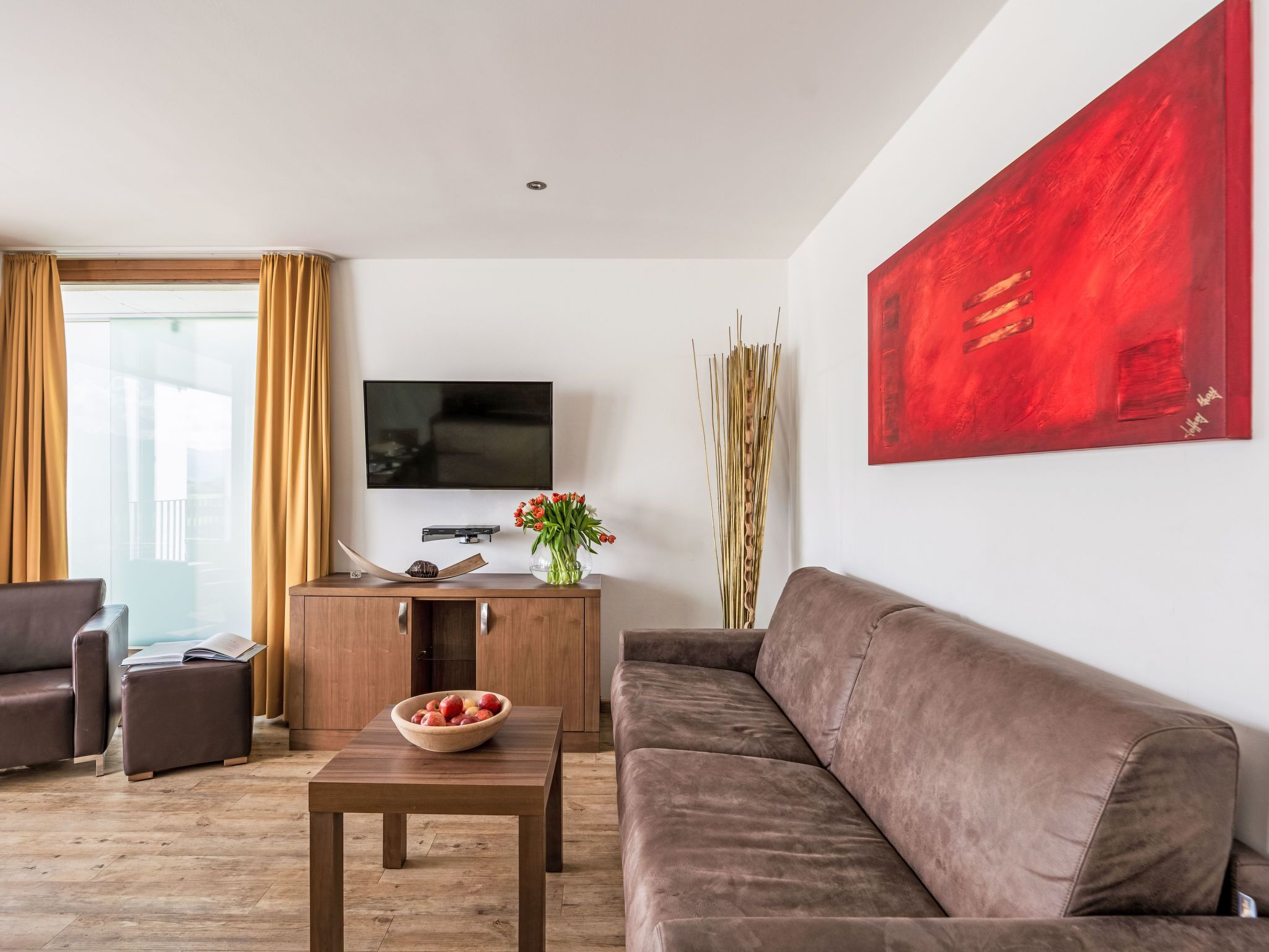 Ein modernes Wohnzimmer mit einem braunen Sofa, einem Holztisch mit einer Fruchtschale, einem Flachbildfernseher auf einer Holzkommode und einem roten, abstrakten Gemälde an der Wand.
