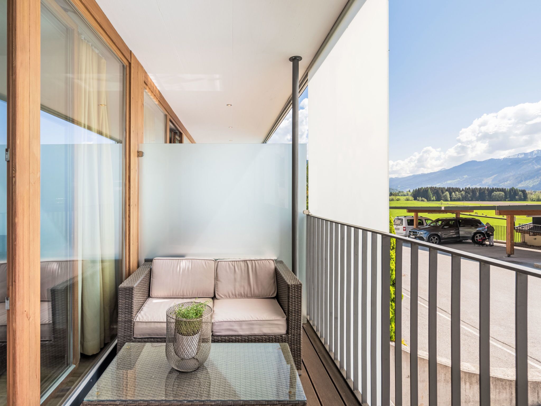 Een balkon met een rieten bank en een glazen tafel, met uitzicht op een parkeerplaats en groene velden.