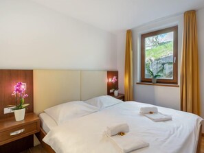 Hotellihuone, jossa on sänky, yöpöytä, ikkuna verhoilla ja näkymä vihreälle alueelle.