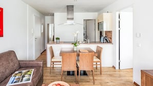 A modern living room with a brown sofa, a dining table with chairs, and a wooden coffee table with apples. There is a kitchen area with a stove, sink, and cabinets in the background. - Apartment Emma by Interhome (Kaprun)