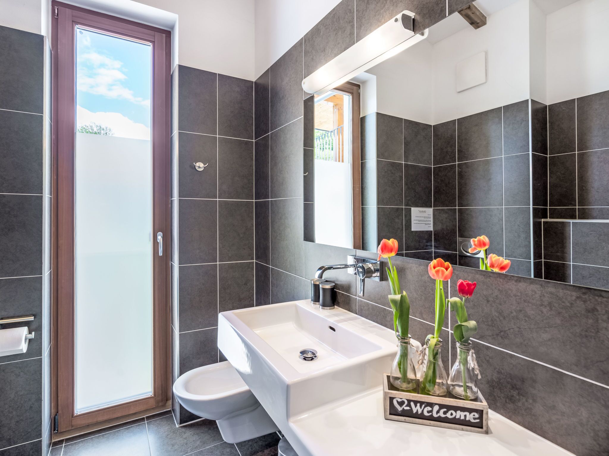 Une salle de bain moderne avec des carreaux foncés, un lavabo blanc, des toilettes et une porte de douche en verre. Un vase rempli de tulipes est posé sur le comptoir du lavabo.