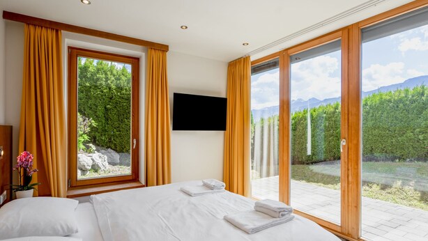 A bedroom with a large bed, a TV mounted on the wall, and a view of a mountainous landscape through large windows. - Apartment Emma by Interhome (Kaprun)