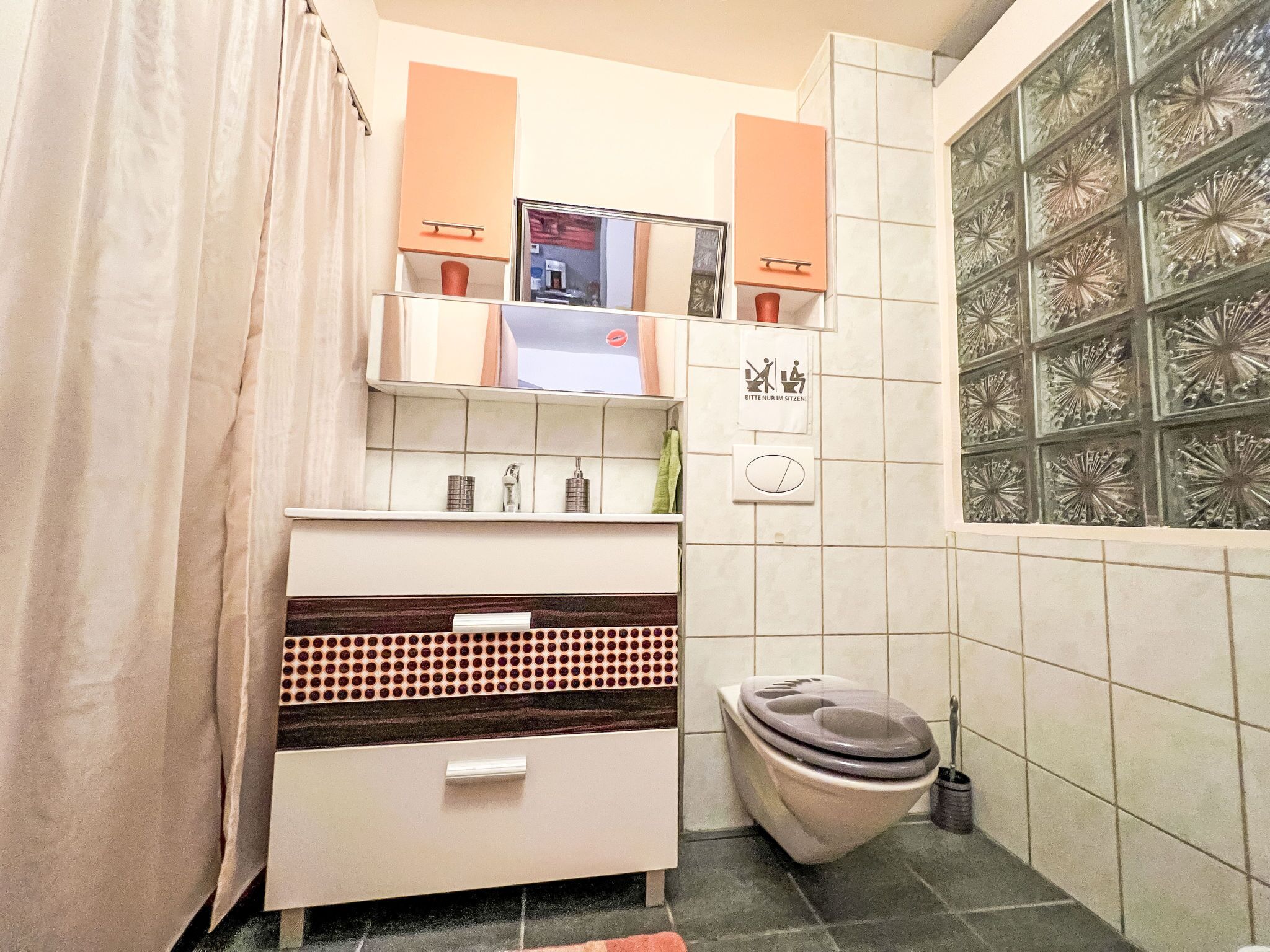 Une salle de bain avec des toilettes, un lavabo et un miroir. Il y a un porte-serviettes au-dessus du lavabo et une poubelle à côté des toilettes.