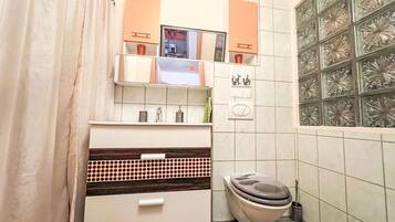 Une salle de bain avec des toilettes, un lavabo et un miroir. Il y a un porte-serviettes au-dessus du lavabo et une poubelle à côté des toilettes.