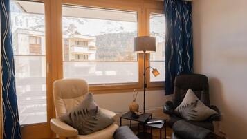 Ein Zimmer mit zwei Ledersesseln, einem kleinen Tisch mit einer Lampe und Blick auf Gebäude durch große Fenster.