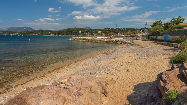 A pebble beach with clear water, a rocky shoreline, and a distant mountain range. - Les Roches Bleues by Interhome (Le Pradet)
