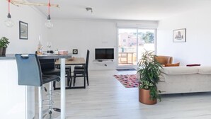 A modern living room with a sofa, a dining area with a table and chairs, a TV, and a large window with a view of buildings. - Les Roches Bleues by Interhome (Le Pradet)