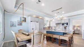 Una cocina amplia con gabinetes blancos, una isla central y un comedor con mesa de madera y sillas de mimbre.