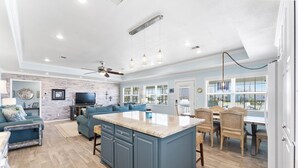 Una cocina amplia con una isla central, comedor con sillas de mimbre y sala de estar con sofá y televisor.
