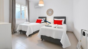 Quarto de hotel com duas camas, roupa de cama branca, almofadas vermelhas, uma luminária pendente e um radiador.