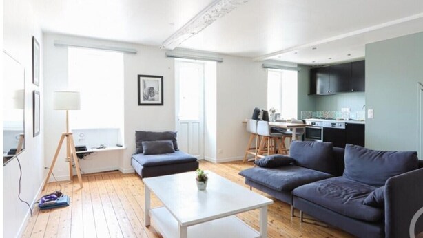A modern living room with a white coffee table, two grey sofas, and a small side table. There is a kitchen area with a TV and cabinets in the background.