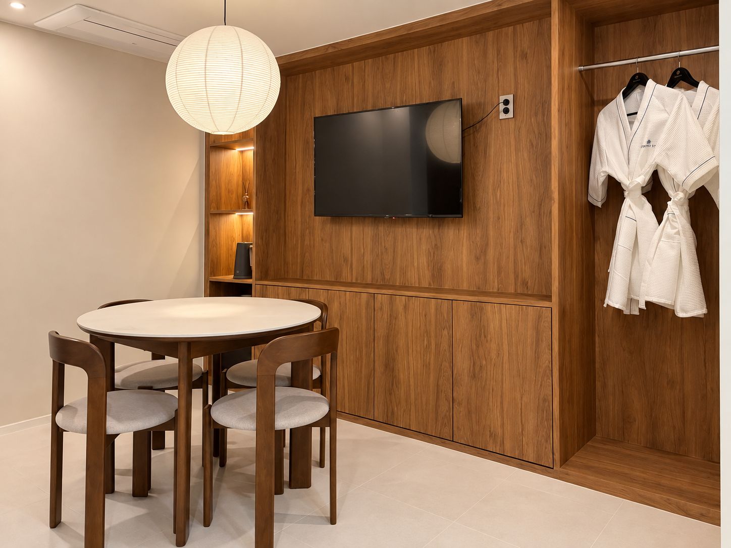 A modern room with a wooden panel wall, a flat-screen TV mounted above a wooden cabinet, a round dining table with chairs, and a clothing rack with white robes.