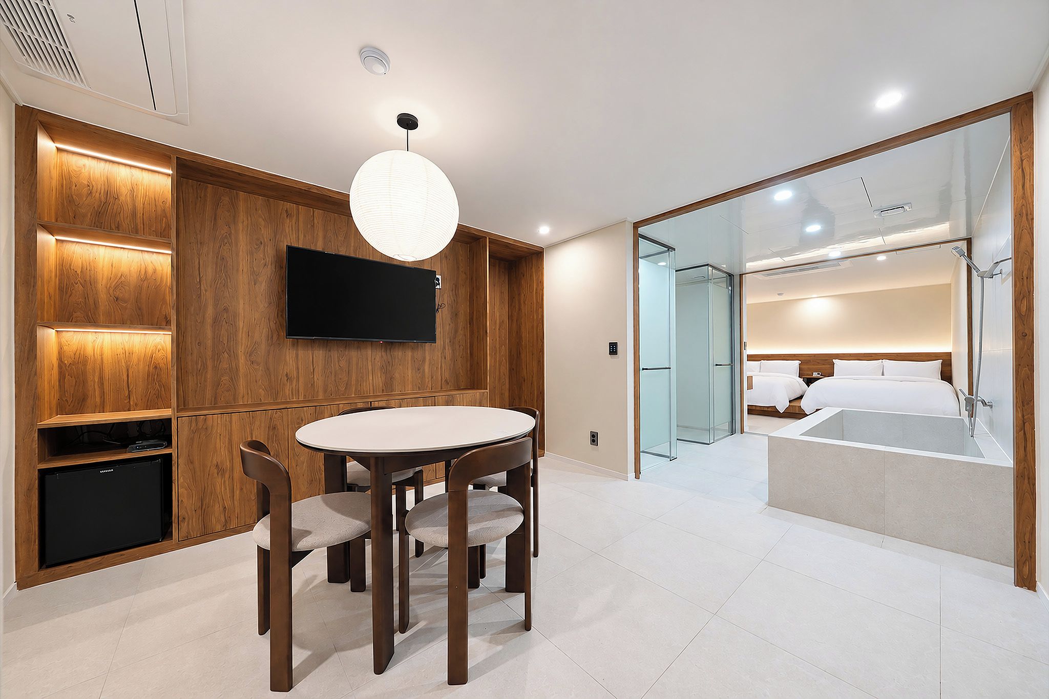 Una moderna camera d'albergo con parete rivestita in legno, televisione a schermo piatto, un tavolo rotondo con sedie e una zona notte separata con un grande letto e cabina doccia in vetro.