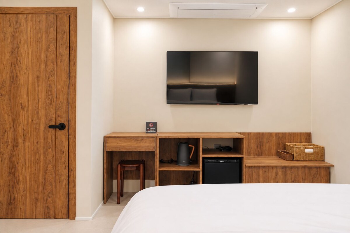 Quarto com cama, escrivaninha de madeira com cadeira, televisão fixada na parede e pequena área de kitchenette.