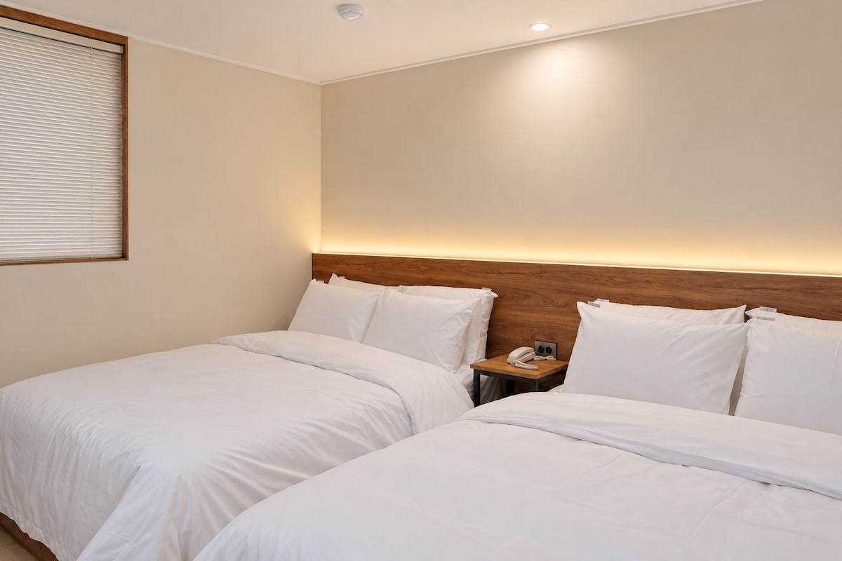 A hotel room with two beds, a wooden headboard, a bedside table with a phone, and a window with blinds.