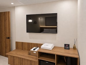 A hotel room with a wooden bench, a flat-screen TV mounted on the wall, a small refrigerator, and a basket with a remote control. - CentroStay (Incheon)
