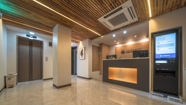 A modern reception area with a wooden ceiling, a reception desk, a digital display, and a trash bin. - CentroStay (Incheon)