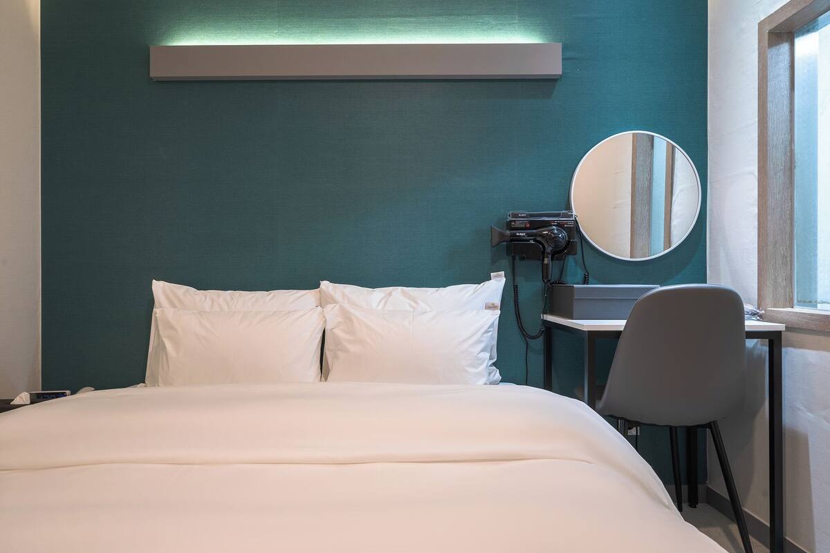 A hotel room with a bed, a desk, a chair, a round mirror, and a window.