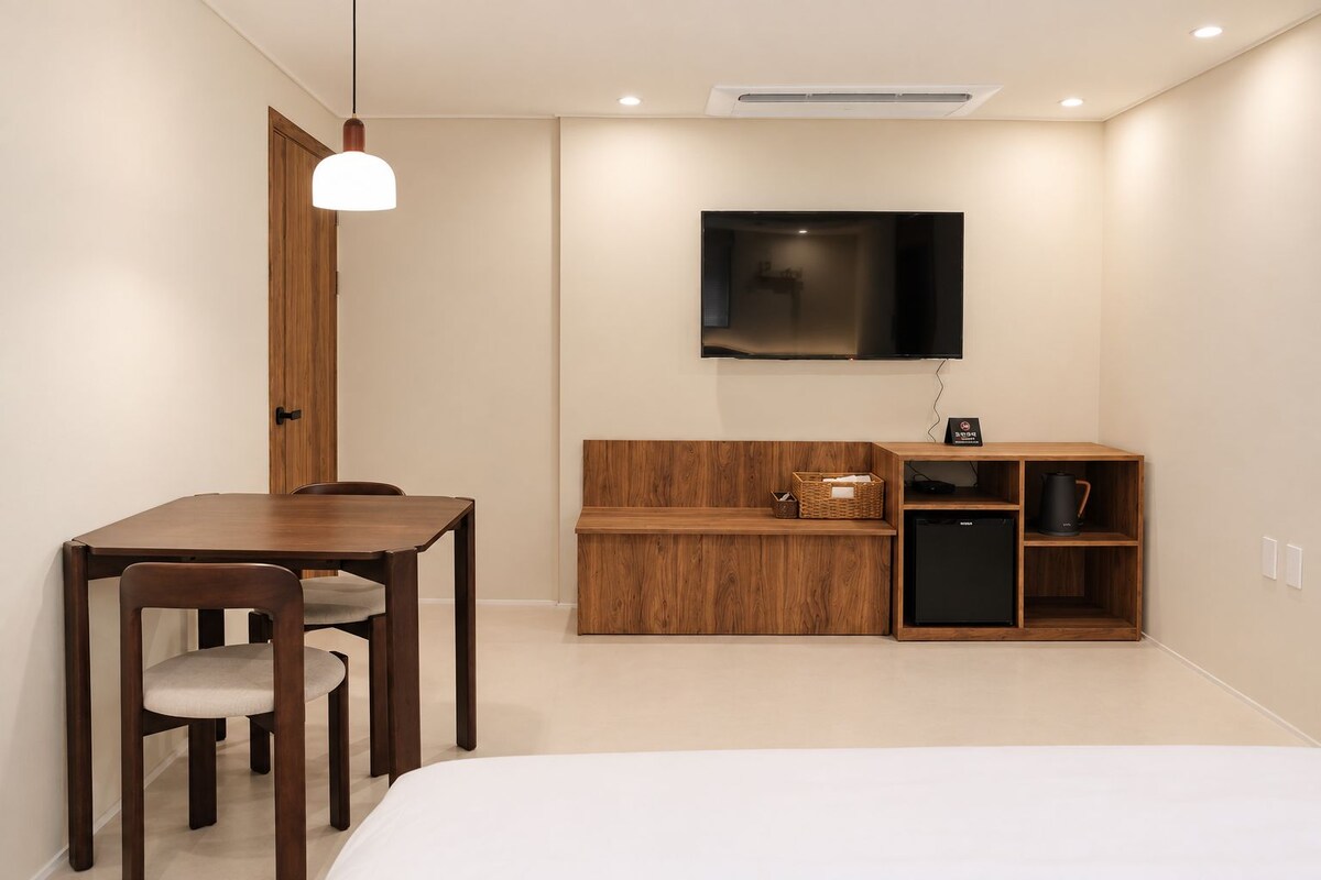 Quarto de hotel moderno com escrivaninha de madeira, uma cadeira, cama, televisão e uma pequena cozinha compacta.