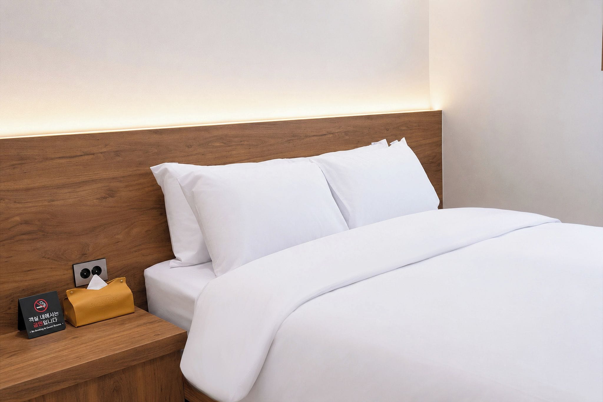 A hotel room with a wooden headboard, white bedding, a bedside table with a tissue box and a no-smoking sign, and a wall-mounted light.