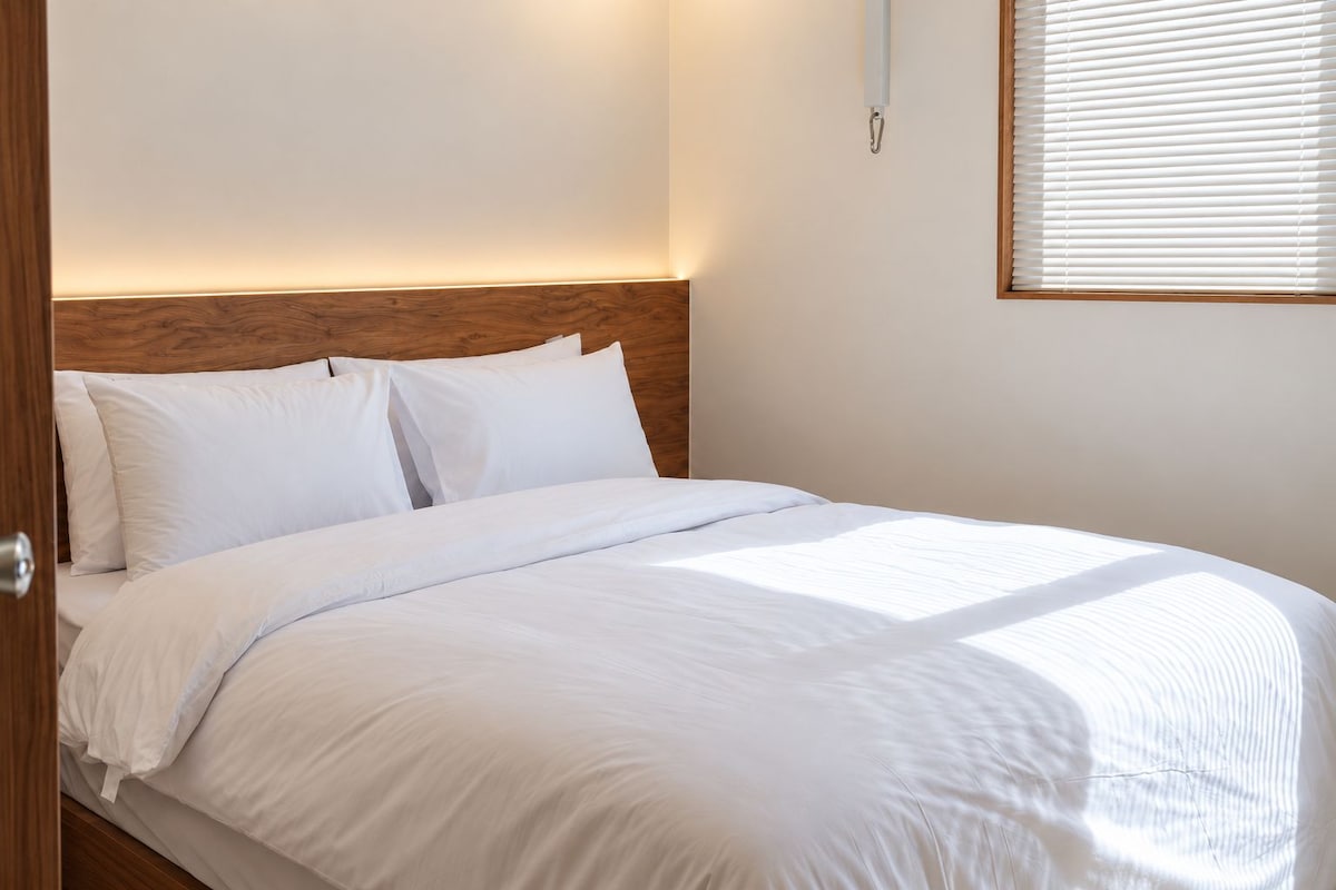 A neatly made bed with white bedding in a room with a wooden headboard and a window with blinds.