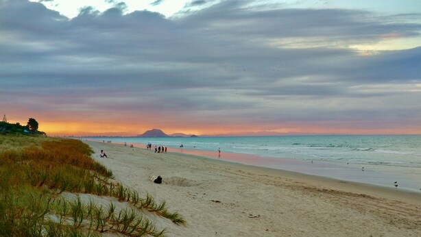 A beach with people walking and standing, a sandy shore, and a sunset in the background. - BACH on BEACH- Pipi Palace. (Papamoa)