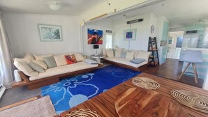A living room with a sofa, a coffee table, and a rug. There is a kitchen area visible in the background. - BACH on BEACH- Pipi Palace. (Papamoa)