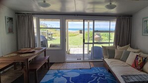 A living room with a sofa, a dining table with benches, and a view of the ocean through large windows. - BACH on BEACH- Pipi Palace. (Papamoa)