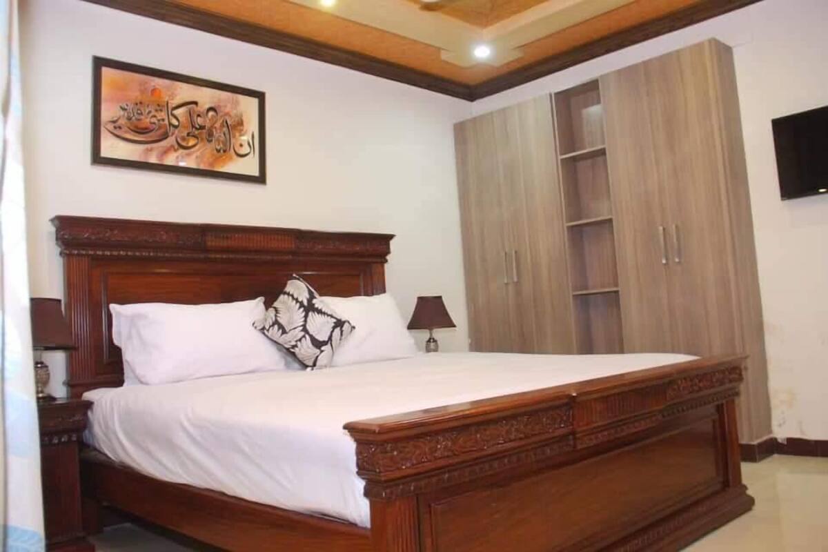 A bedroom with a wooden bed, a nightstand, a lamp, a wardrobe, and a wall-mounted TV.