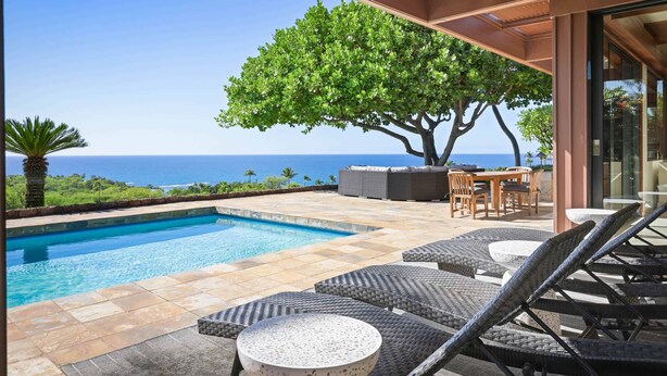 Ein Poolbereich mit Liegestühlen und einem Tisch, ein Baum sowie ein Blick aufs Meer.