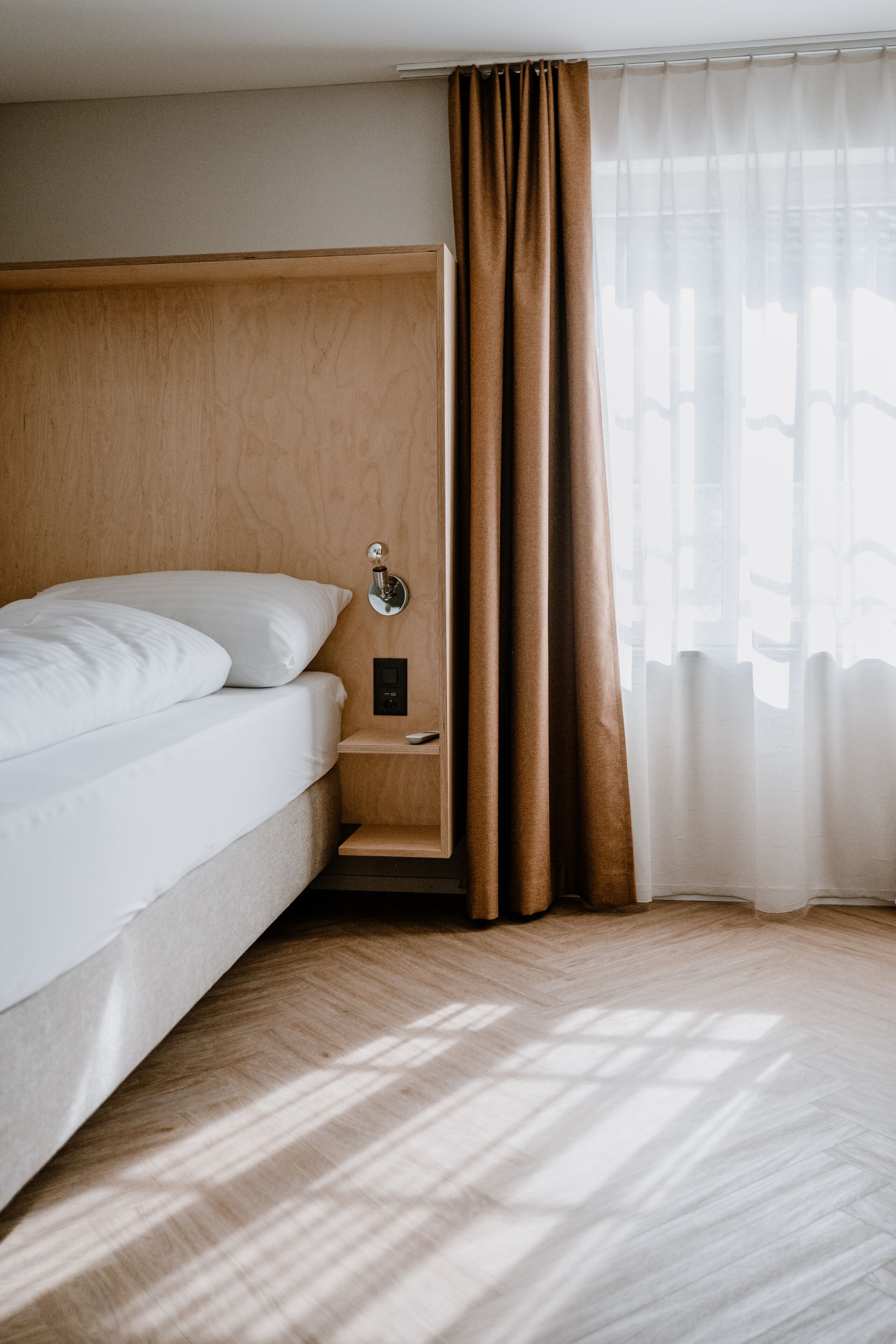 A modern hotel room with a wooden headboard, a single bed with white linens, a bedside table with a lamp, and sheer curtains.