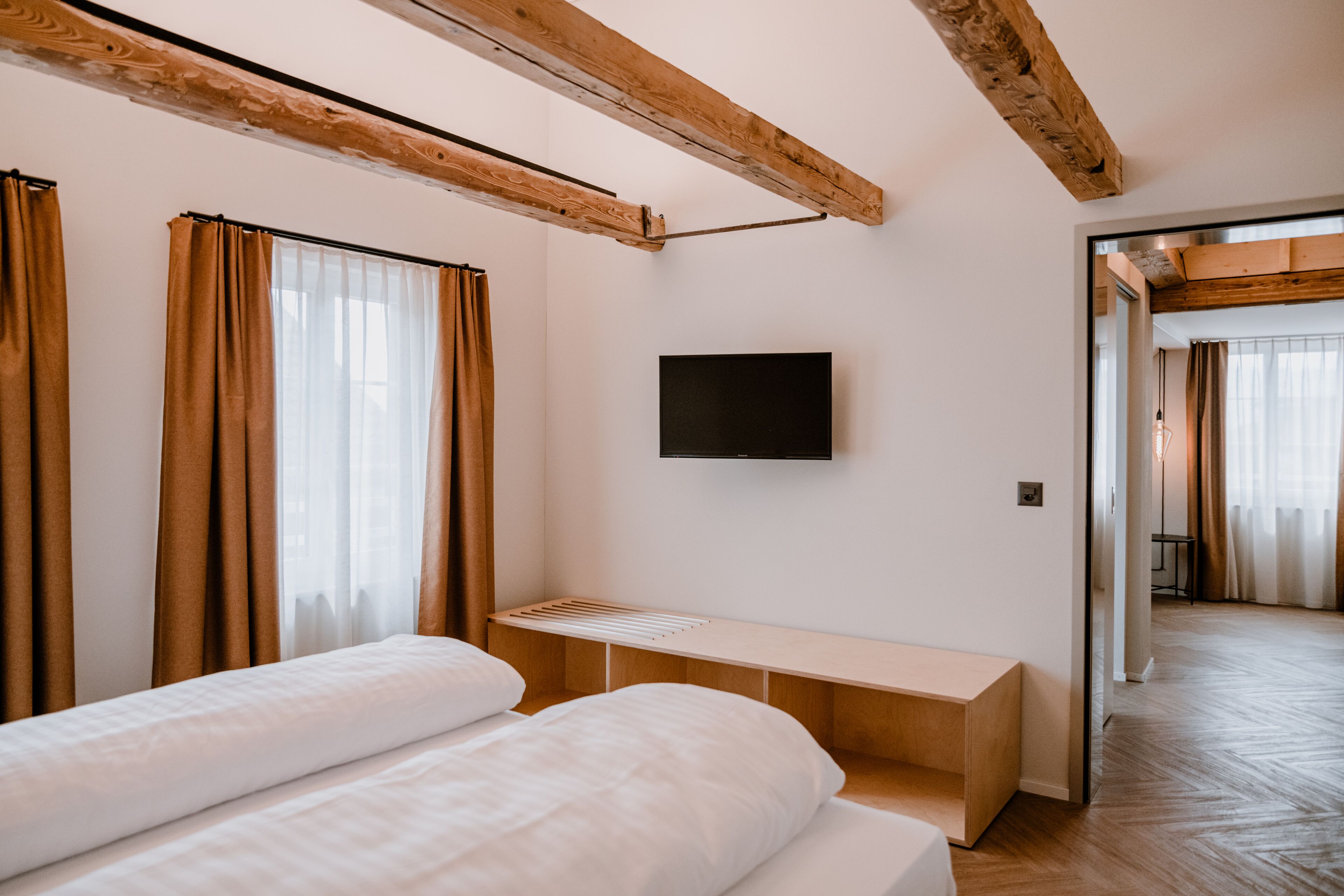 A modern hotel room with a wooden ceiling, a flat-screen TV mounted on the wall, a bed with white linens, and a wooden bench.