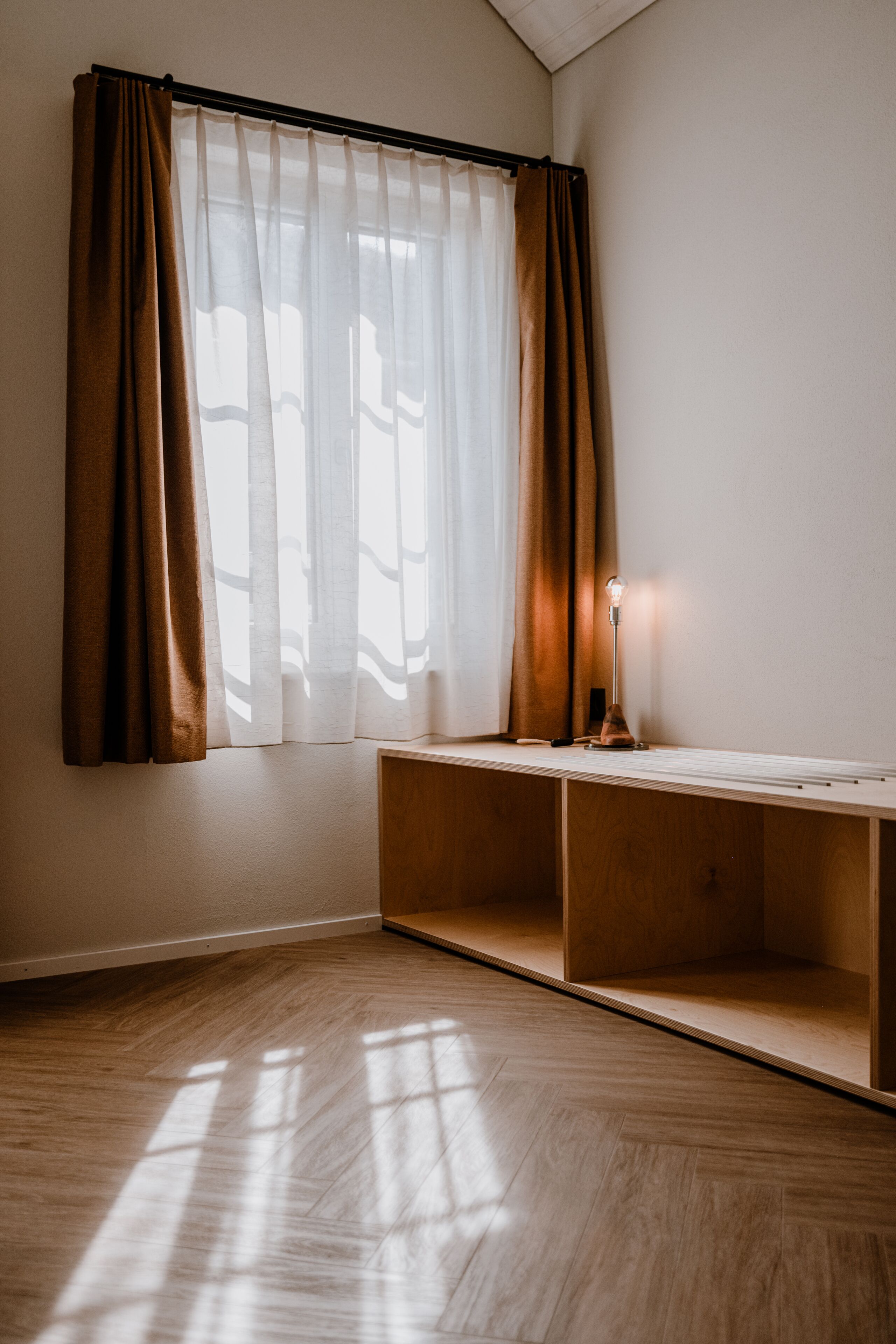 Ein Zimmer mit Holzboden, einem Fenster mit durchsichtigen Vorhängen und einem Schreibtisch mit Lampe.